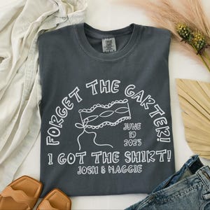May include: Dark gray t-shirt with white text that reads "FORGET THE GARTER! I GOT THE SHIRT!" with the date "JUNE 19 2025" and names "JOSH & MAGGIE". The shirt is surrounded by clothing and accessories.