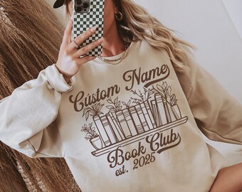 May include: Beige sweatshirt with a brown graphic of a bookshelf, plants, and the text "Custom Name Book Club est. 2025". The sweatshirt has long sleeves and a crew neck.