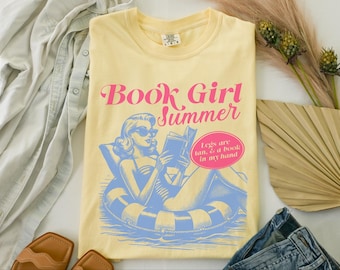 May include: A yellow t-shirt with a pink and blue graphic of a woman reading a book in a pool float. The text on the shirt reads "Book Girl Summer" and "Legs are tan, & a book in my hand."