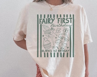 May include: Cream-colored t-shirt with a green and white graphic that reads "FAIRY FIRST Birthday" and "OLIVIA'S 1ST BIRTHDAY." The graphic includes a fairy, cupcake, mushrooms, butterflies, and musical notes. The shirt is a relaxed fit.