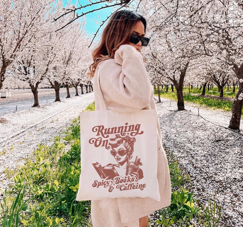 May include: A white tote bag with a brown graphic design featuring a woman reading a book and the text "Running on Spicy Books & Caffeine".