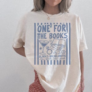 May include: Cream-colored t-shirt with a blue graphic that reads "ONE FOR THE BOOKS" above a martini glass, ring, and book illustration. Below, it says "OLIVIA'S BACHELORETTE New York, New York".