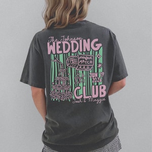 Op de afbeelding: Donkergrijs t-shirt met een roze en groen grafisch ontwerp op de achterkant. Het ontwerp bevat de woorden "The Johnson WEDDING CLUB" en de datum "JUN 19 2026". Extra afbeeldingen zijn champagneglazen en muzieknoten.
