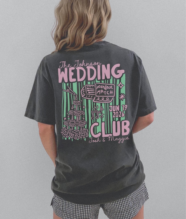 Op de afbeelding: Donkergrijs t-shirt met een roze en groen grafisch ontwerp op de achterkant. Het ontwerp bevat de woorden "The Johnson WEDDING CLUB" en de datum "JUN 19 2026". Extra afbeeldingen zijn champagneglazen en muzieknoten.