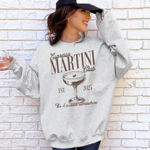 May include: A light gray sweatshirt with the text "Espresso MARTINI Club" in brown, along with a martini glass graphic. The sweatshirt also includes the text "EST. 2025" and "It's 5 o'clock somewhere."