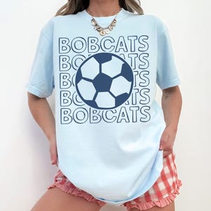 K&ouml;nnte beinhalten: Ein hellblaues Kurzarm-T-Shirt mit einer dunkelblauen Fu&szlig;ballgrafik in der Mitte der Brust. Das Wort BOBCATS ist in einem sich wiederholenden, umrandeten Schriftmuster hinter der Fu&szlig;ballgrafik in Dunkelblau aufgedruckt.
