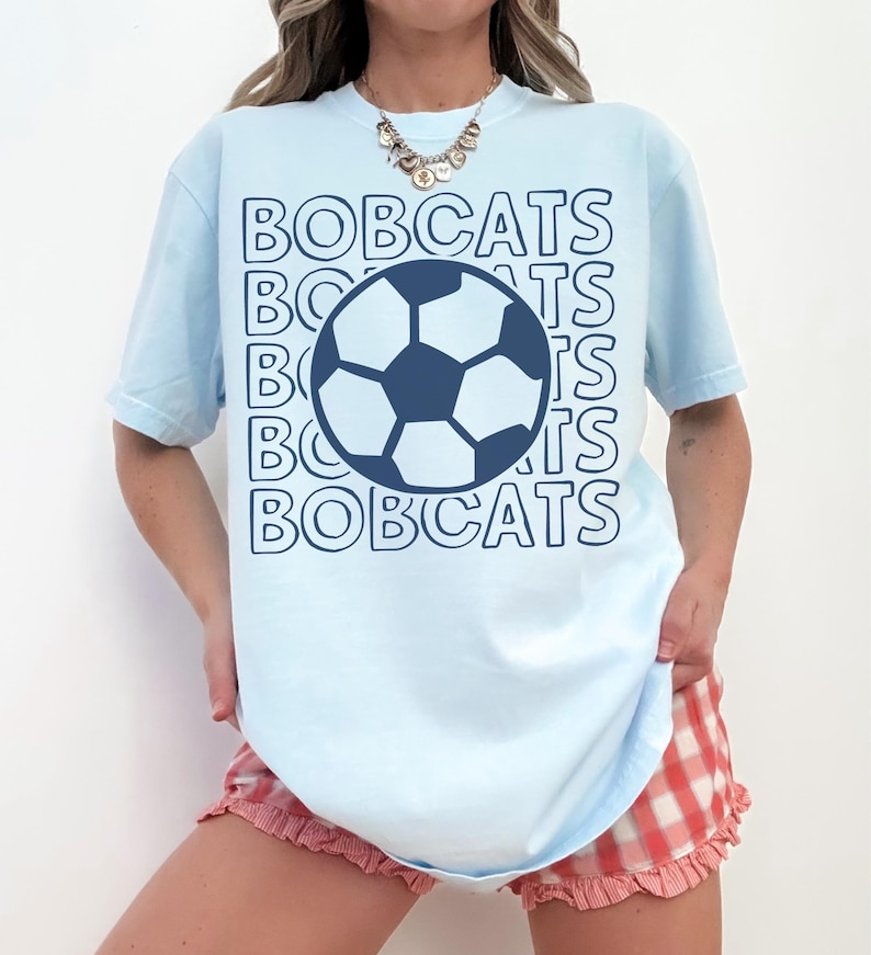 K&ouml;nnte beinhalten: Ein hellblaues Kurzarm-T-Shirt mit einer dunkelblauen Fu&szlig;ballgrafik in der Mitte der Brust. Das Wort BOBCATS ist in einem sich wiederholenden, umrandeten Schriftmuster hinter der Fu&szlig;ballgrafik in Dunkelblau aufgedruckt.