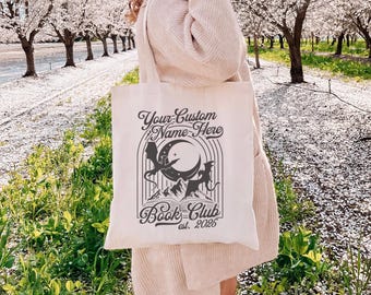 Puede incluir: Bolso tote color crema con un diseño gráfico de un dragón, una luna y montañas. El texto en el bolso dice "Your Custom Name Here" y "Book Club est. 2025". El bolso se lleva al aire libre.