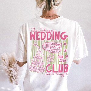 Op de afbeelding: Cr&egrave;mekleurig T-shirt met een roze en groen grafisch ontwerp. Het ontwerp bevat de tekst "The Johnson WEDDING CLUB" samen met illustraties van champagneglazen, muzieknoten en de datum "JUN 19 2026".