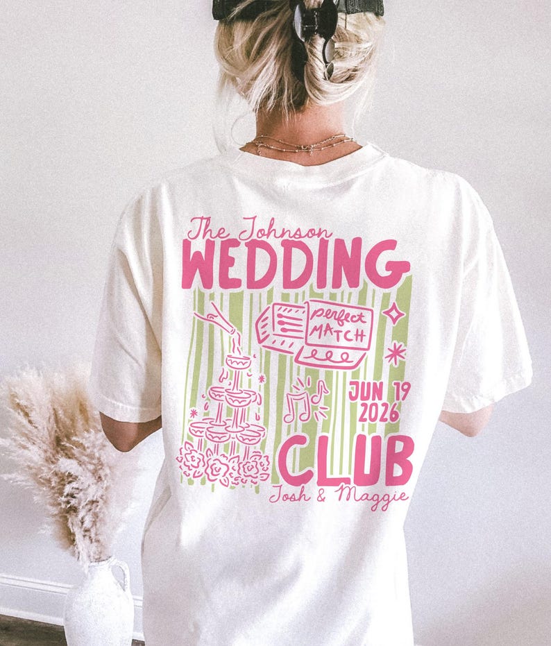 Op de afbeelding: Cr&egrave;mekleurig T-shirt met een roze en groen grafisch ontwerp. Het ontwerp bevat de tekst "The Johnson WEDDING CLUB" samen met illustraties van champagneglazen, muzieknoten en de datum "JUN 19 2026".
