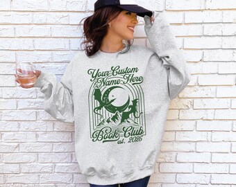 May include: A light gray sweatshirt with a green graphic design featuring a crescent moon, dragons, and mountains. The text "Your Custom Name Here" and "Book Club est. 2025" are also printed on the sweatshirt.