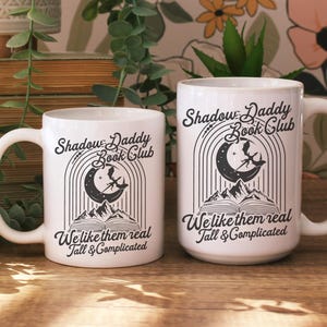 May include: Two white ceramic mugs with black text and illustrations. The mugs feature the text "Shadow Daddy Book Club" and "We like them real Tall & Complicated".