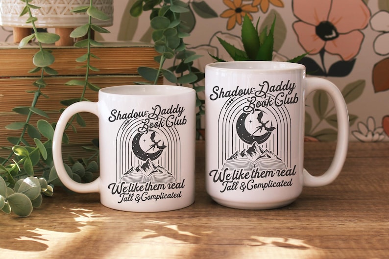 May include: Two white ceramic mugs with black text and illustrations. The mugs feature the text "Shadow Daddy Book Club" and "We like them real Tall & Complicated".