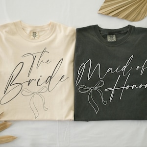 May include: Two t-shirts, one cream and one dark gray, with script text and bow graphics. The cream shirt reads "The Bride," and the dark gray shirt reads "Maid of Honor." Both shirts are folded and displayed on a light surface.