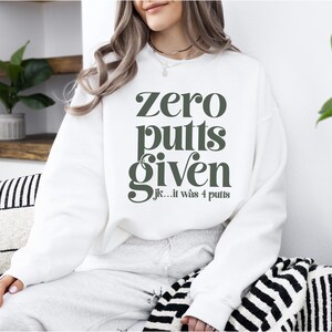 May include: White sweatshirt with the text "zero putts given jk... it was 4 putts" in green.