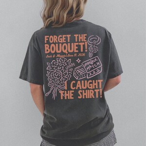 May include: Dark gray t-shirt with the text "FORGET THE BOUQUET!" in orange, along with the names "Josh & Maggie" and the date "June 19, 2026". The shirt also features a bouquet graphic and the phrase "I CAUGHT THE SHIRT!"