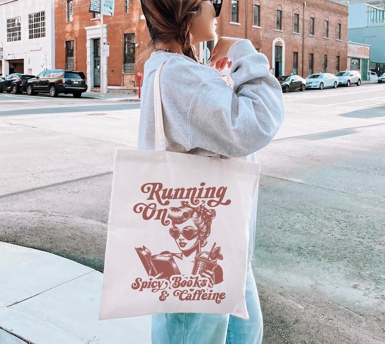 May include: White canvas tote bag with a brown graphic of a woman reading a book and holding a drink. The text on the bag reads "Running on Spicy Books & Caffeine".