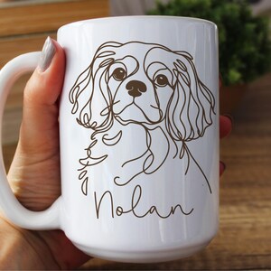 May include: White ceramic mug featuring a line drawing of a Cavalier King Charles Spaniel dog. The dog's image is in brown, with the name "Nolan" in a script font below. The mug has a large handle.