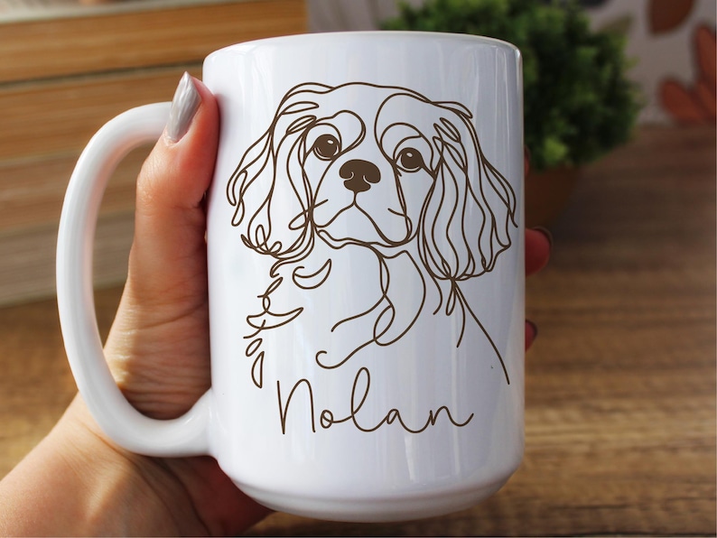 May include: White ceramic mug featuring a line drawing of a Cavalier King Charles Spaniel dog. The dog's image is in brown, with the name "Nolan" in a script font below. The mug has a large handle.