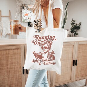 May include: White tote bag with a red graphic of a woman reading a book and holding a drink. The text on the bag reads "Running on Spicy Books & Caffeine".