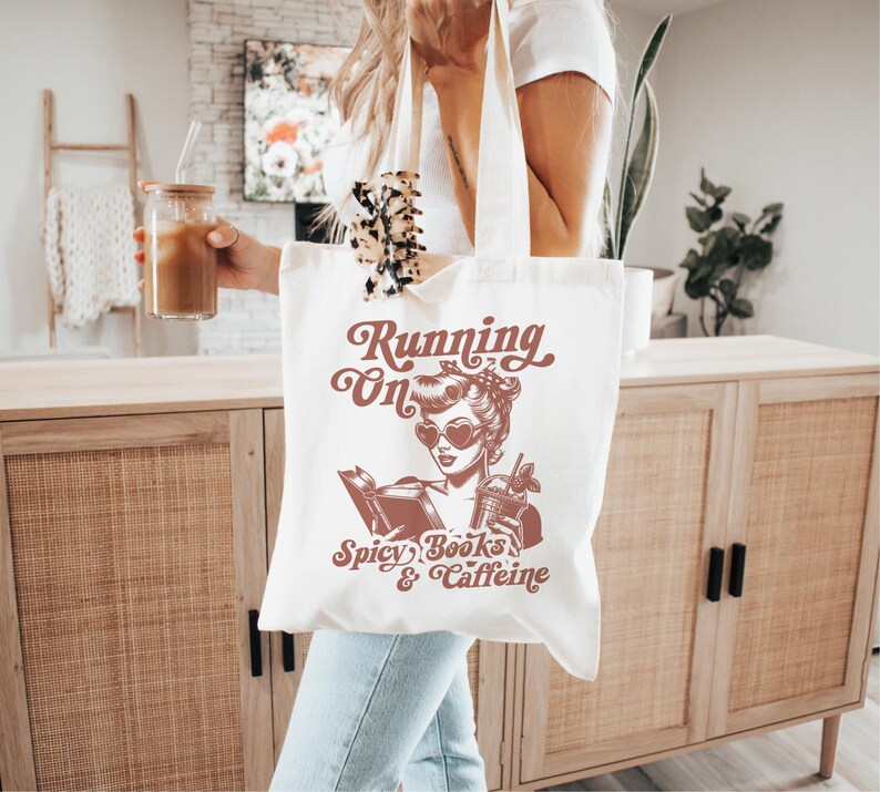 May include: White tote bag with a red graphic of a woman reading a book and holding a drink. The text on the bag reads "Running on Spicy Books & Caffeine".