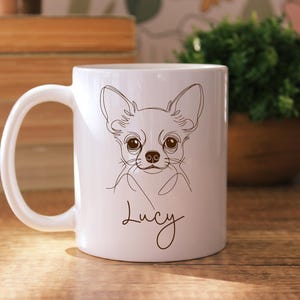 May include: White ceramic mug featuring a line drawing of a Chihuahua dog with large eyes and ears. The dog's name, "Lucy", is written in a cursive font below the drawing. The mug has a curved handle and is set against a wooden surface.