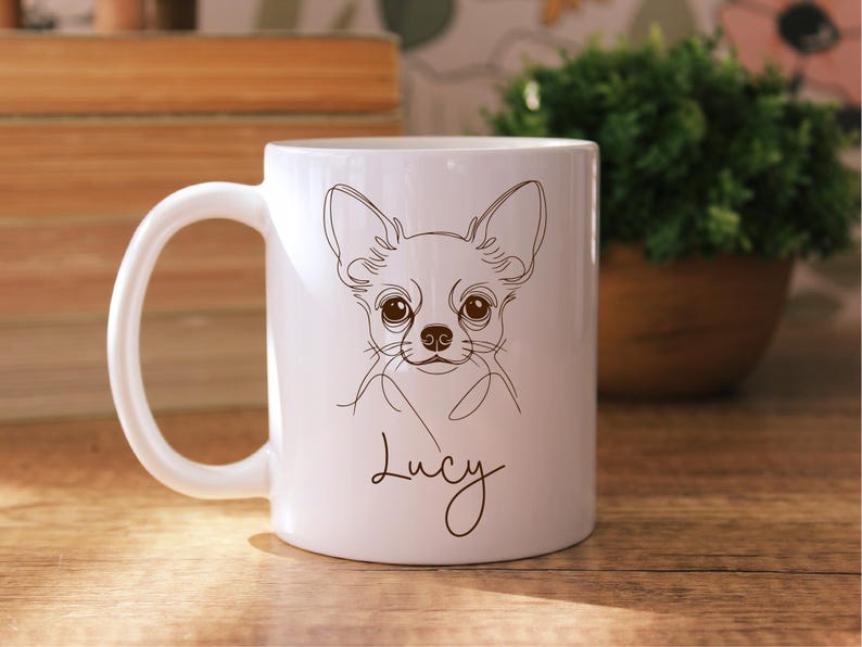 May include: White ceramic mug featuring a line drawing of a Chihuahua dog with large eyes and ears. The dog's name, "Lucy", is written in a cursive font below the drawing. The mug has a curved handle and is set against a wooden surface.