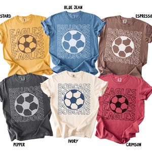 K&ouml;nnte beinhalten: Sechs kurz&auml;rmelige T-Shirts in den Farben Senf, Jeansblau, Espresso, Pfeffer, Elfenbein und Karmesinrot. Jedes T-Shirt zeigt eine zentrale Fu&szlig;ballgrafik, umgeben von wiederholten Teamnamen wie Eagles, Bulldogs und Bobcats in wei&szlig;er oder schwarzer Umriss-Schrift.