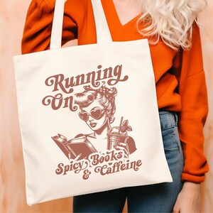 May include: A white tote bag with a red design featuring a woman with a book and a drink. The text on the bag reads "Running on Spicy Books & Caffeine".