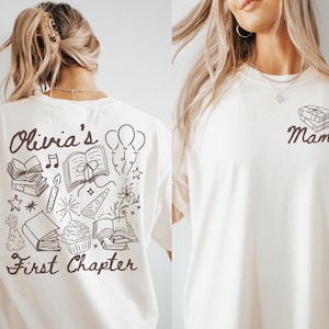May include: Cream-colored t-shirt with a black graphic. The back features the text "Olivia's First Chapter" with illustrations of books, balloons, and a cupcake. The front has the word "Mama" with a stack of books.