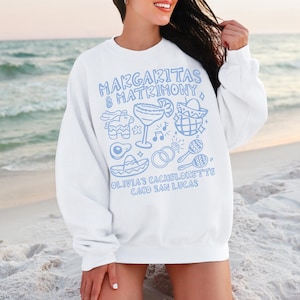 May include: White sweatshirt with light blue graphic text and illustrations. The text reads "Margaritas & Matrimony" and "Olivia's Bachelorette Cabo San Lucas." The illustrations include a margarita, a piñata, and maracas.