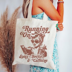 May include: A white canvas tote bag with a brown graphic of a woman wearing heart-shaped sunglasses and holding a book and a drink. The text on the bag reads "Running on Spicy Books & Caffeine".