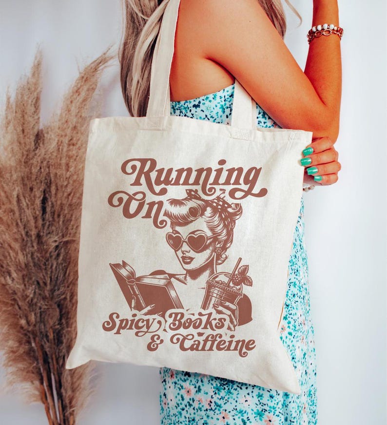 May include: A white canvas tote bag with a brown graphic of a woman wearing heart-shaped sunglasses and holding a book and a drink. The text on the bag reads "Running on Spicy Books & Caffeine".