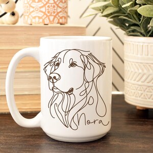 May include: White ceramic mug featuring a line drawing of a dog's face in brown ink. The dog's name, "Nora", is written in a cursive font below the drawing. The mug has a large handle and is suitable for hot or cold beverages.