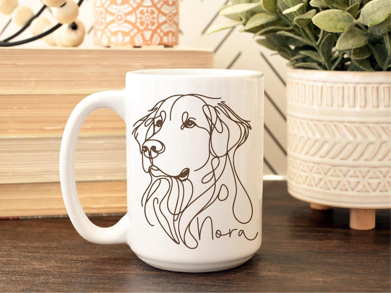 May include: White ceramic mug featuring a line drawing of a dog's face in brown ink. The dog's name, "Nora", is written in a cursive font below the drawing. The mug has a large handle and is suitable for hot or cold beverages.