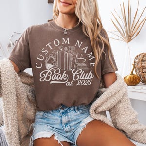 May include: A brown t-shirt with a white graphic that reads "CUSTOM NAME Book Club est. 2025". The graphic includes illustrations of books, flowers, and a coffee cup. The person is wearing denim shorts and a cream-colored sweater.