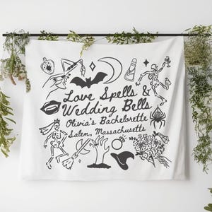 May include: White fabric wall hanging with black illustrations and text. The text reads "Love Spells & Wedding Bells Olivia's Bachelorette Salem, Massachusetts." The design includes a bat, moon, potion bottle, and skeleton.