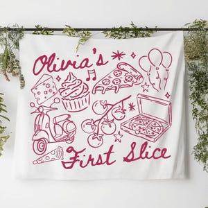 May include: White fabric wall hanging featuring a burgundy illustration of pizza, a cupcake, a scooter, and other food items. The text "Olivia's First Slice" is written in a cursive font.