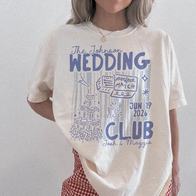 Op de afbeelding: Cr&egrave;mekleurig T-shirt met een blauw grafisch ontwerp. Het ontwerp bevat de woorden "The Johnson WEDDING CLUB" en de datum "JUN 19 2026". Extra elementen zijn een champagnetoren, muzieknoten en de woorden "perfect MATCH".