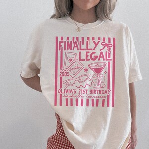 May include: A cream-coloured t-shirt featuring a pink graphic. The graphic includes the text "FINALLY LEGAL" and illustrations of a heart, sunglasses, a cocktail, and "OLIVIA'S 21ST BIRTHDAY Nashville, Tennessee".