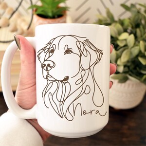 May include: White ceramic mug featuring a continuous line drawing of a dog's face in brown. The dog's name, "Nora", is written in a cursive font below the drawing. The mug has a large handle.