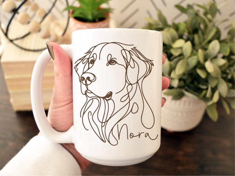 May include: White ceramic mug featuring a continuous line drawing of a dog's face in brown. The dog's name, "Nora", is written in a cursive font below the drawing. The mug has a large handle.