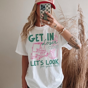 May include: White t-shirt with a golf-themed graphic. The design features the text "GET IN Loser, LET'S LOOK for your ball" in green and pink, along with illustrations of a golf cart and golf bag. The shirt is a casual, oversized fit.