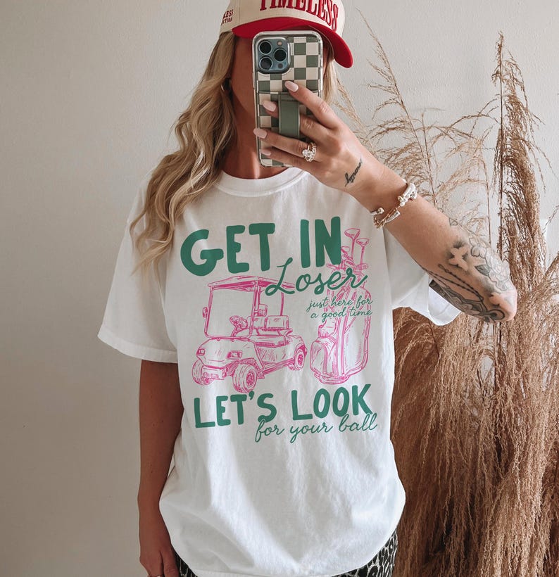 May include: White t-shirt with a golf-themed graphic. The design features the text "GET IN Loser, LET'S LOOK for your ball" in green and pink, along with illustrations of a golf cart and golf bag. The shirt is a casual, oversized fit.