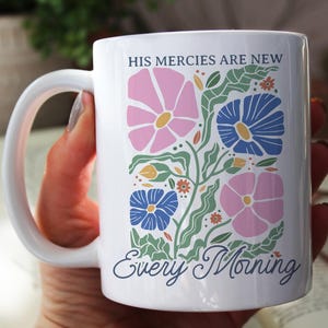 K&ouml;nnte beinhalten: Wei&szlig;e Keramiktasse mit einem Blumendesign in Rosa, Blau und Gr&uuml;n. Die Tasse tr&auml;gt den Text "HIS MERCIES ARE NEW Every Morning" in einer Schreibschrift. Die Tasse hat einen gro&szlig;en, gebogenen Henkel.