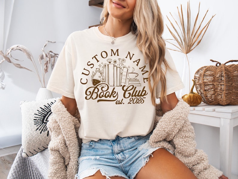 May include: Cream-colored t-shirt with a brown graphic that reads "CUSTOM NAME Book Club est. 2025." The design includes illustrations of books, a coffee cup, and a potion bottle. The shirt is worn with denim shorts.
