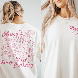 May include: Cream-colored t-shirt with pink text and illustrations. The back features the words "Olivia's Berry First Birthday" with strawberry and party-themed graphics. The front has the word "Mama" and a strawberry.