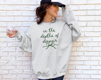 May include: Light gray sweatshirt with the text "in the depths of despair" in dark green script. The sweatshirt has long sleeves and a crew neck. A green bow graphic is below the text. The person in the image is wearing a black and white baseball cap.
