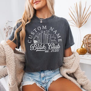 May include: Dark gray t-shirt with a white graphic that reads "CUSTOM NAME Book Club est. 2025." The design includes books, flowers, and a coffee cup. The model is wearing denim shorts and a cream-colored jacket.