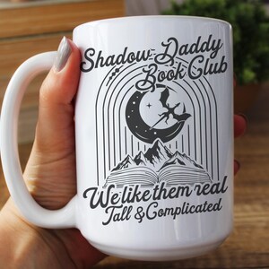 May include: White ceramic mug with a black graphic design featuring a crescent moon, a dragon, and a book. The text reads "Shadow Daddy Book Club We like them real Tall & Complicated".